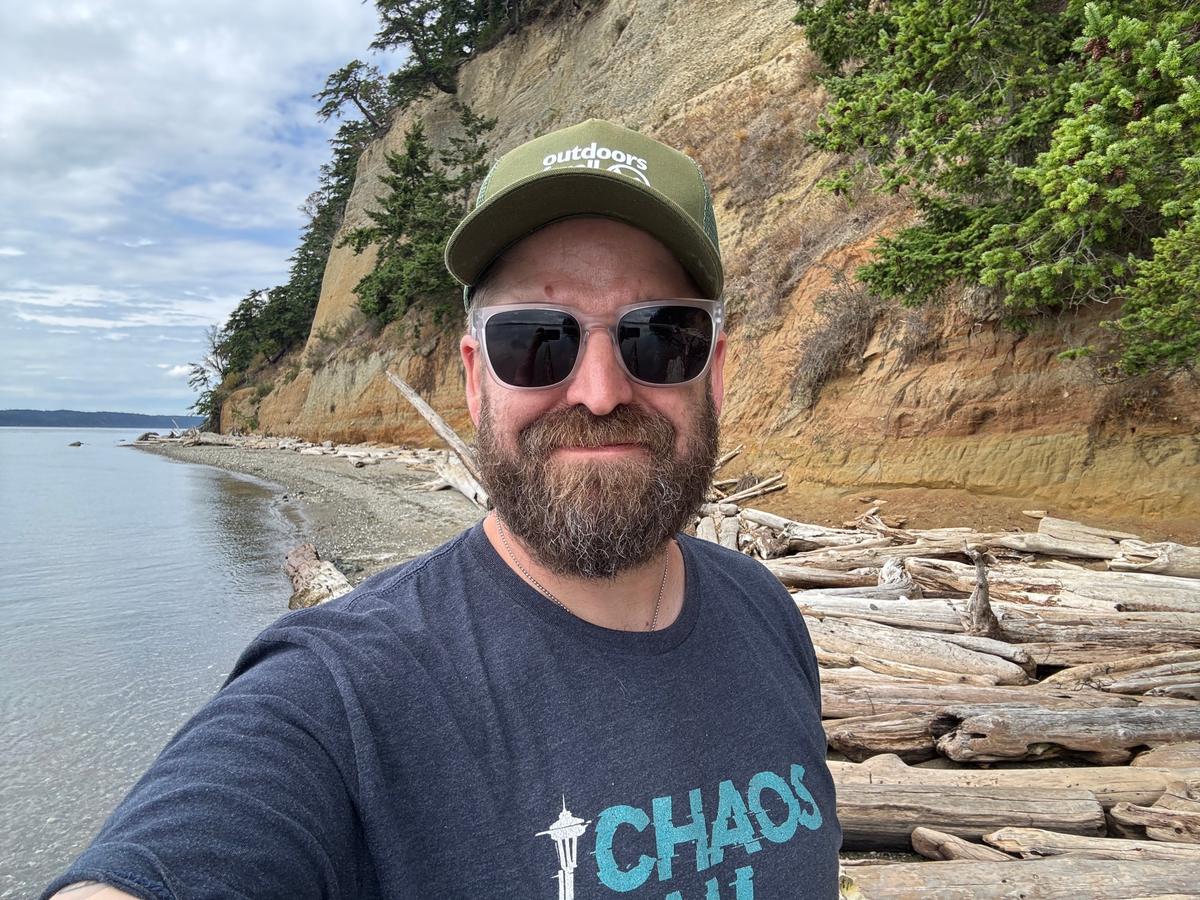 Matt Lyman smiling in sunglasses and a green cap on a driftwood beach below a sandy bluff