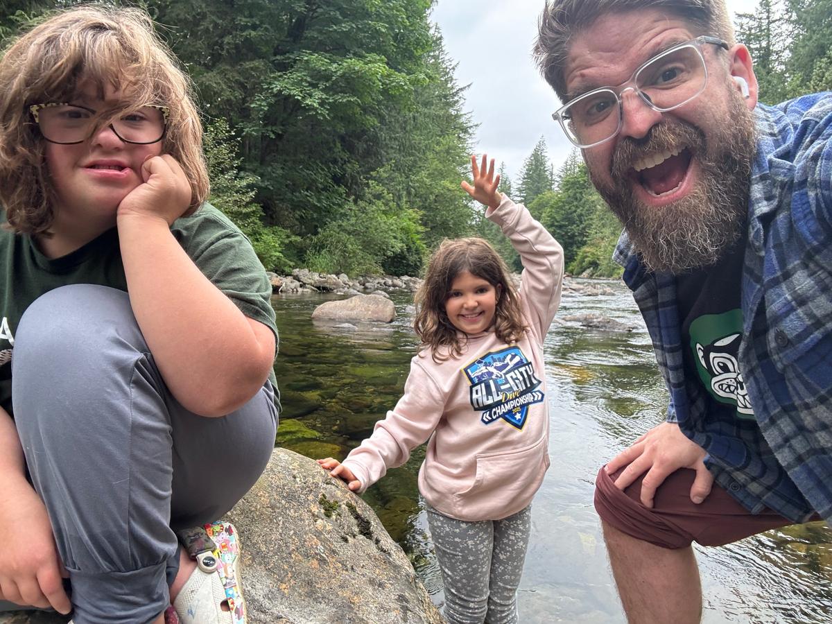 Matt Lyman making a big open-mouthed grin in a selfie with two kids beside a river